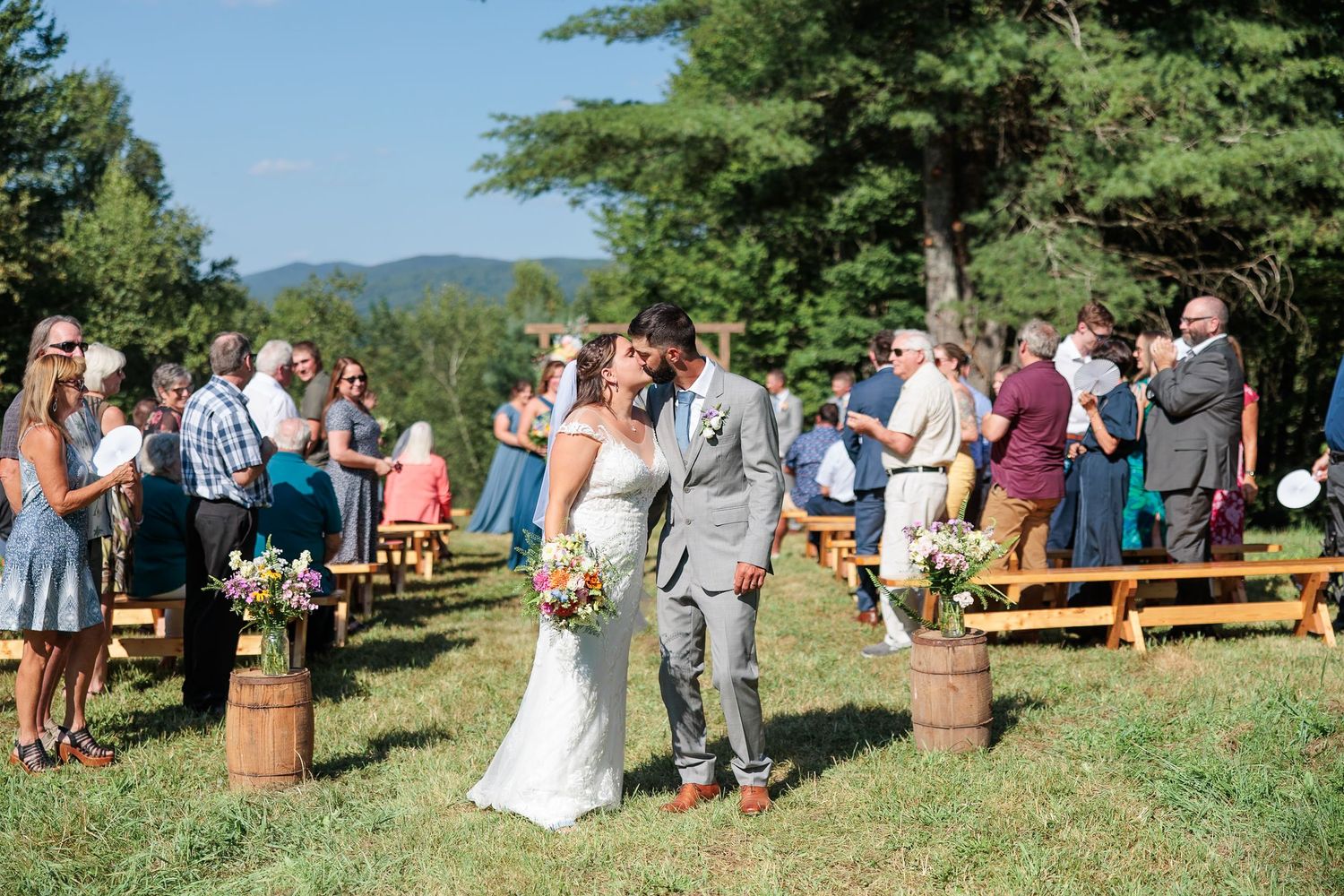 Bride and groom share first kiss at outdoor wedding ceremony surrounded by guests and mountain views.