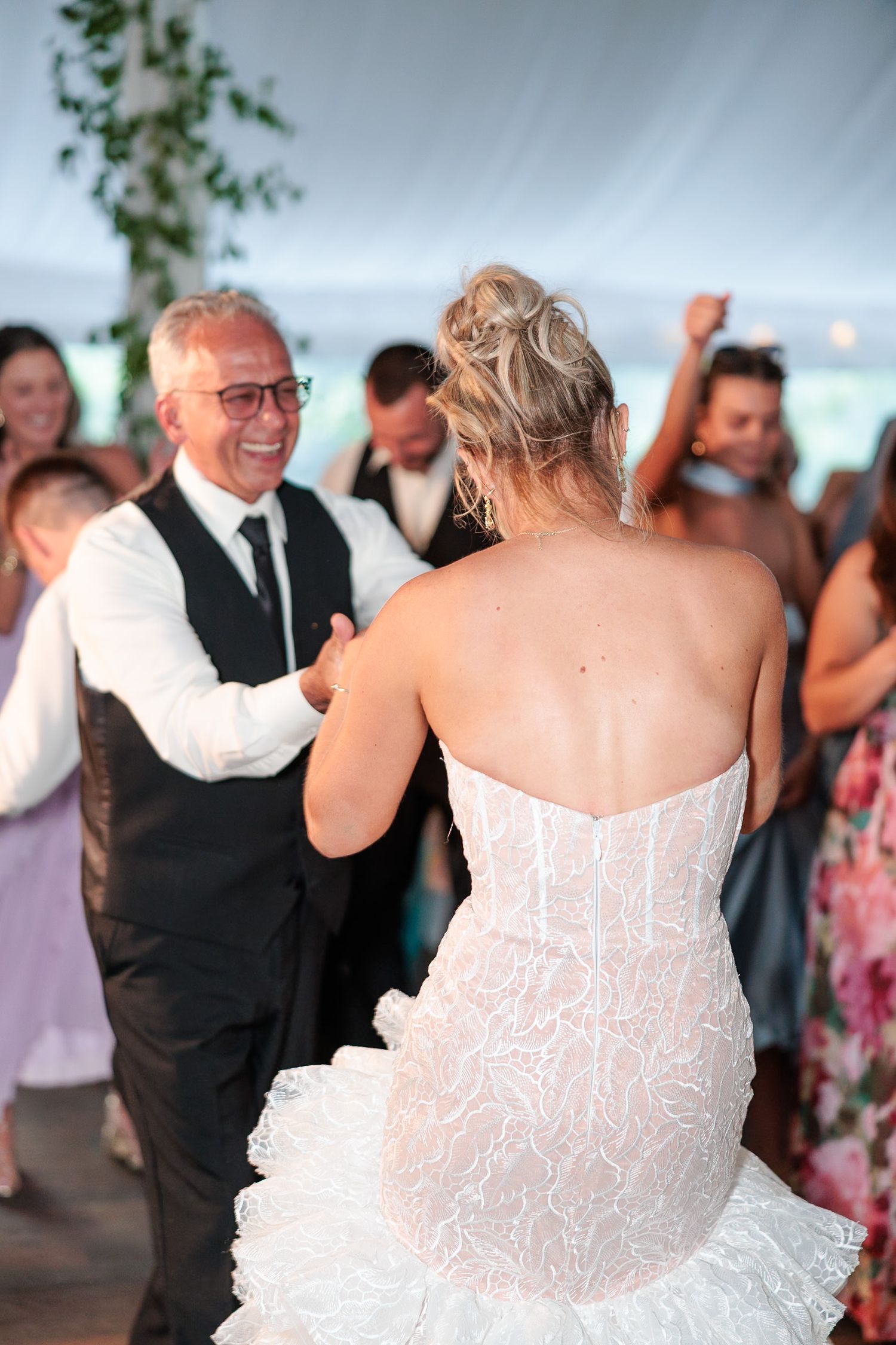 Bride in strapless lace mermaid gown dances with older man in black suit at elegant tented wedding reception.
