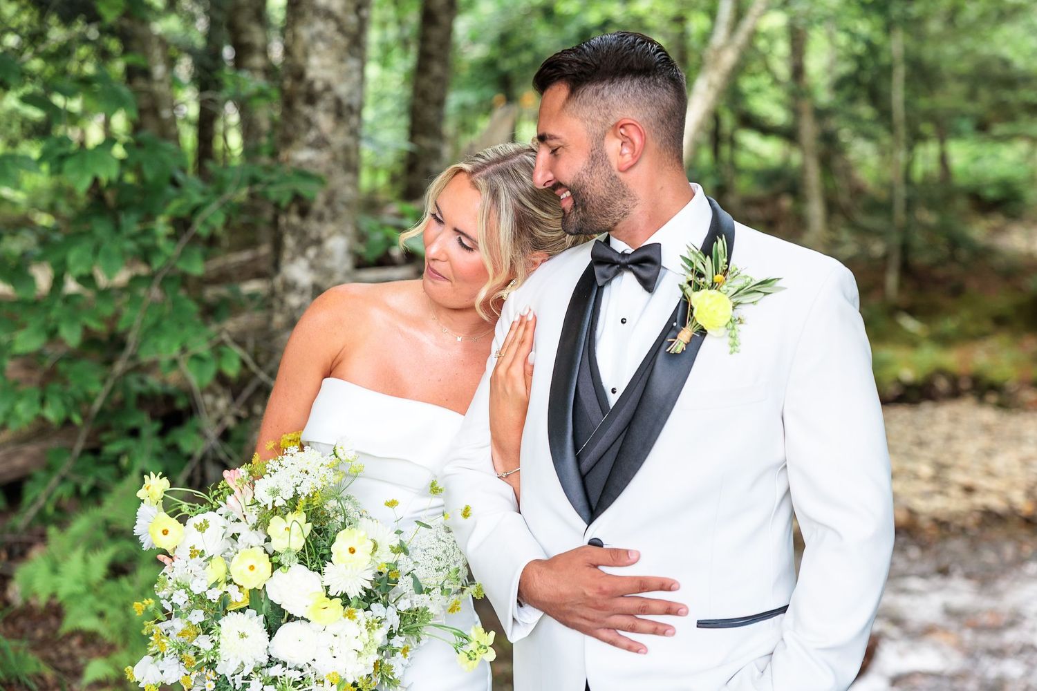 Bride and groom share a romantic moment in a lush forest setting, groom in white tuxedo with yellow boutonniere.