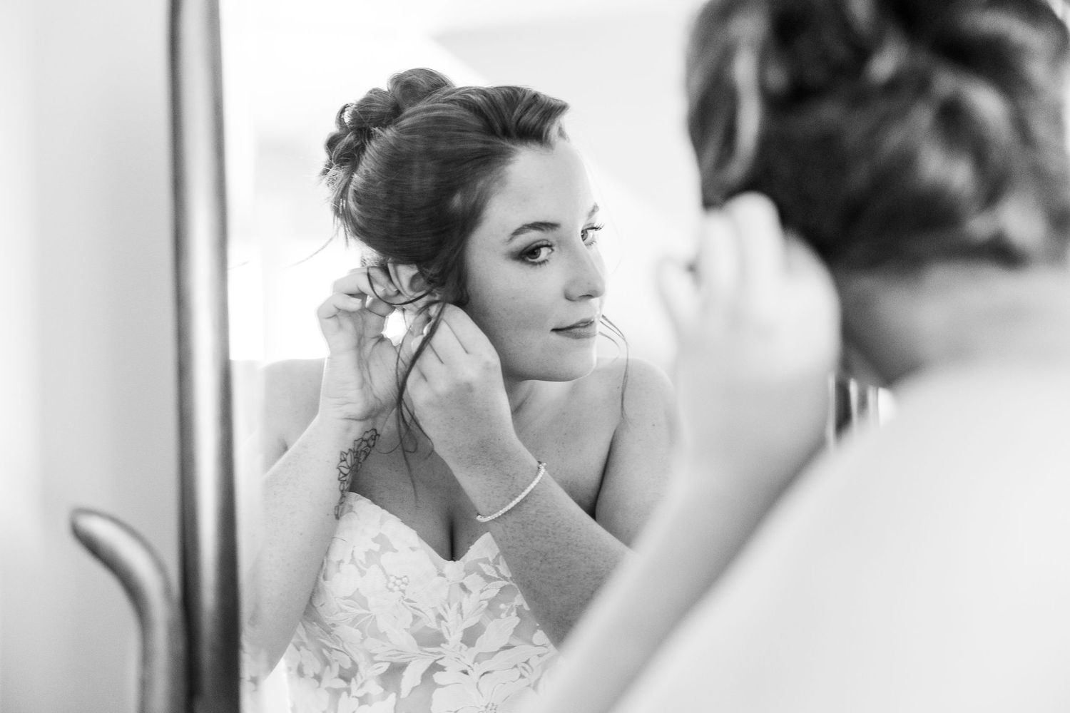 Bride putting on earrings while looking in mirror in black and white wedding photo.