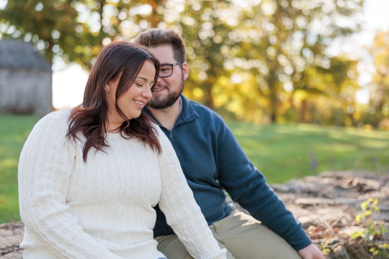Engaged couple sitting closely together outdoors in warm golden sunlight during fall season.