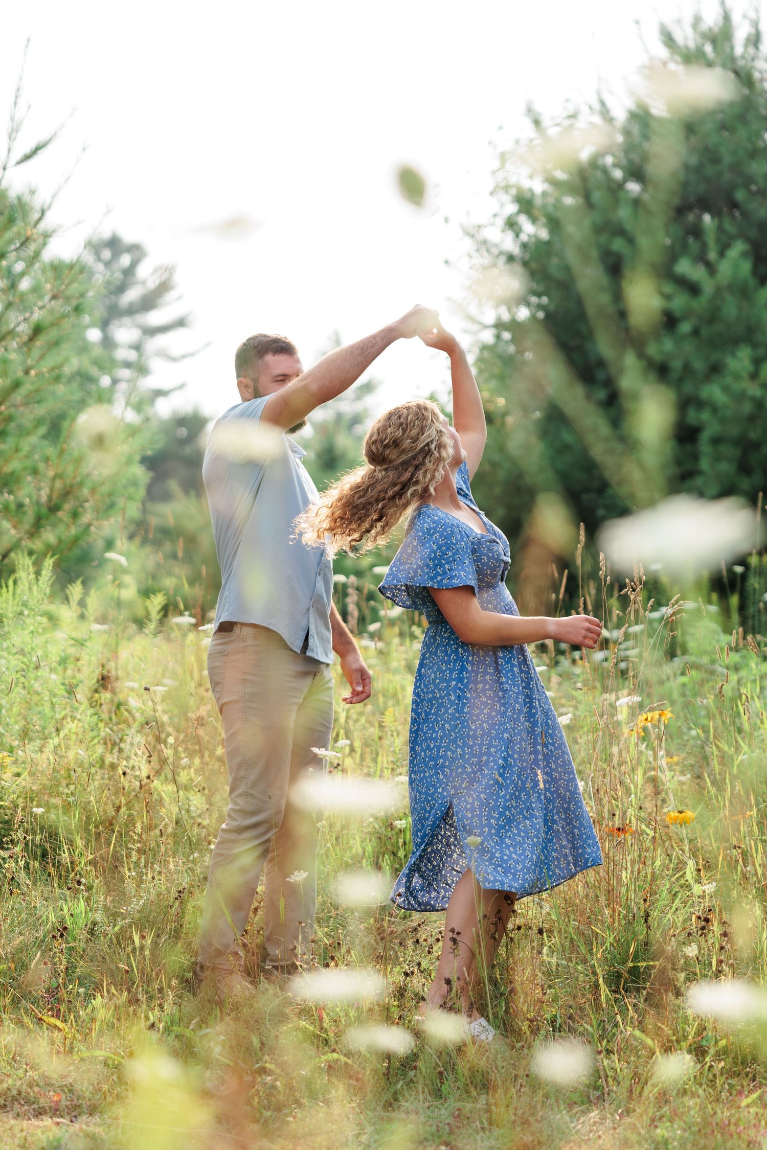 Man spinning woman in blue floral dress through a sunlit wildflower meadow on a summer day.