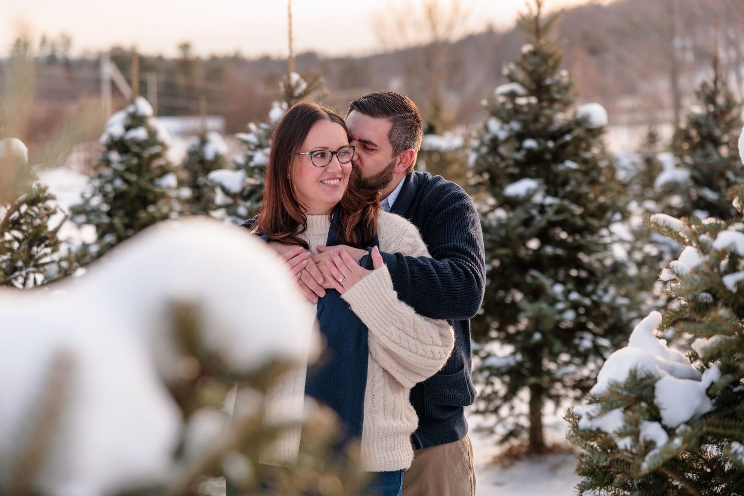 Man hugging woman from behind among snow-dusted evergreen trees at sunset.