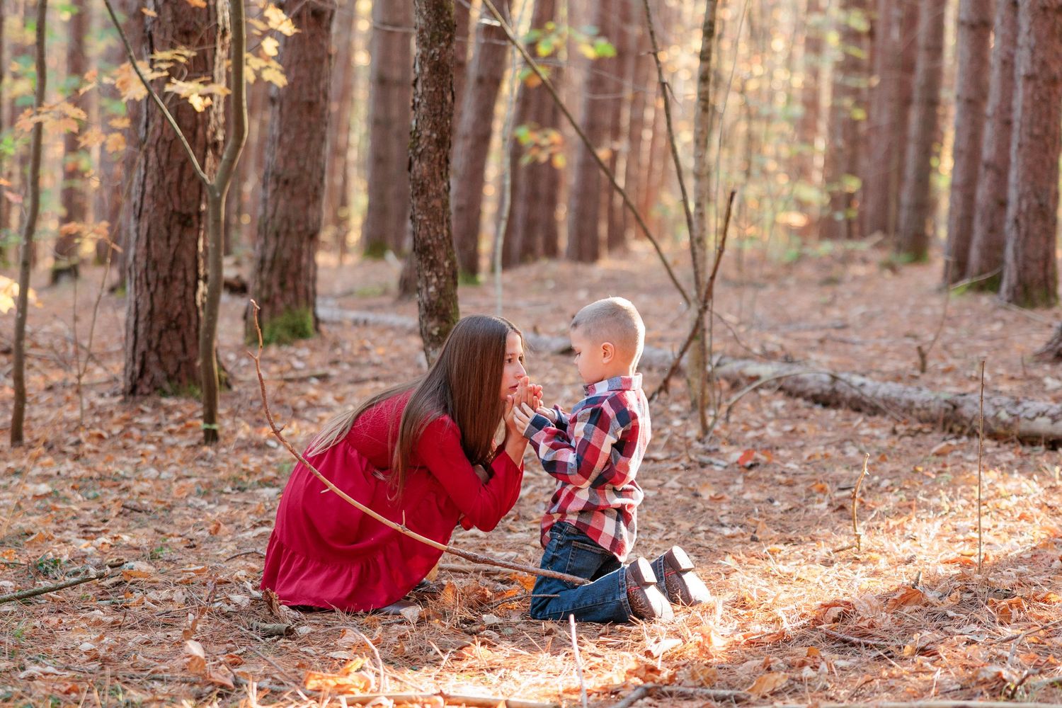 Mother in red dress kneeling to interact with toddler son in plaid shirt among autumn forest trees.