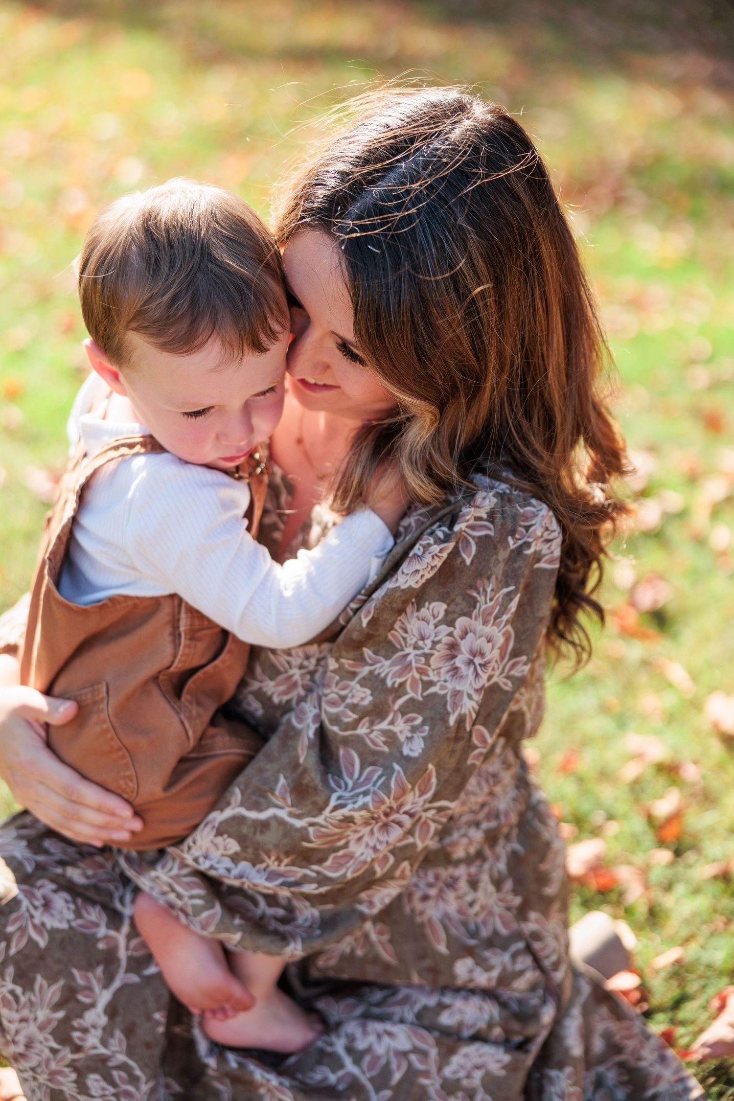 Mother in floral dress holding and cuddling smiling toddler in brown overalls on autumn lawn.
