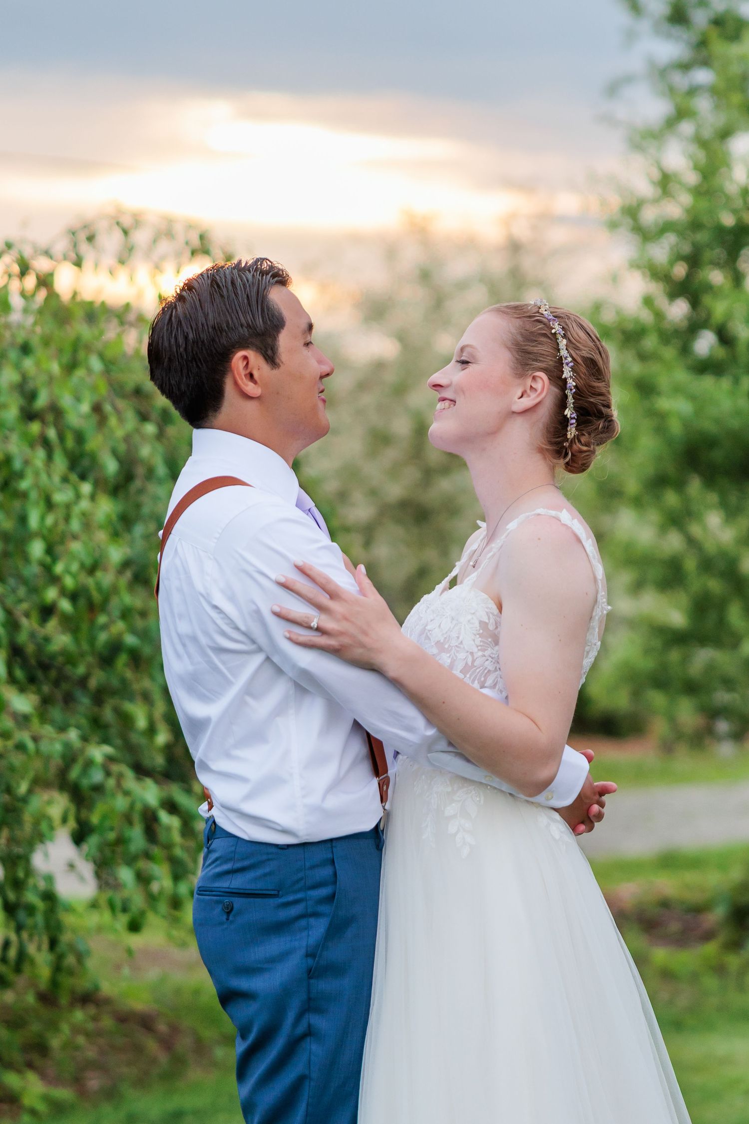 Bride and groom gaze lovingly at each other outdoors at sunset surrounded by lush green trees.