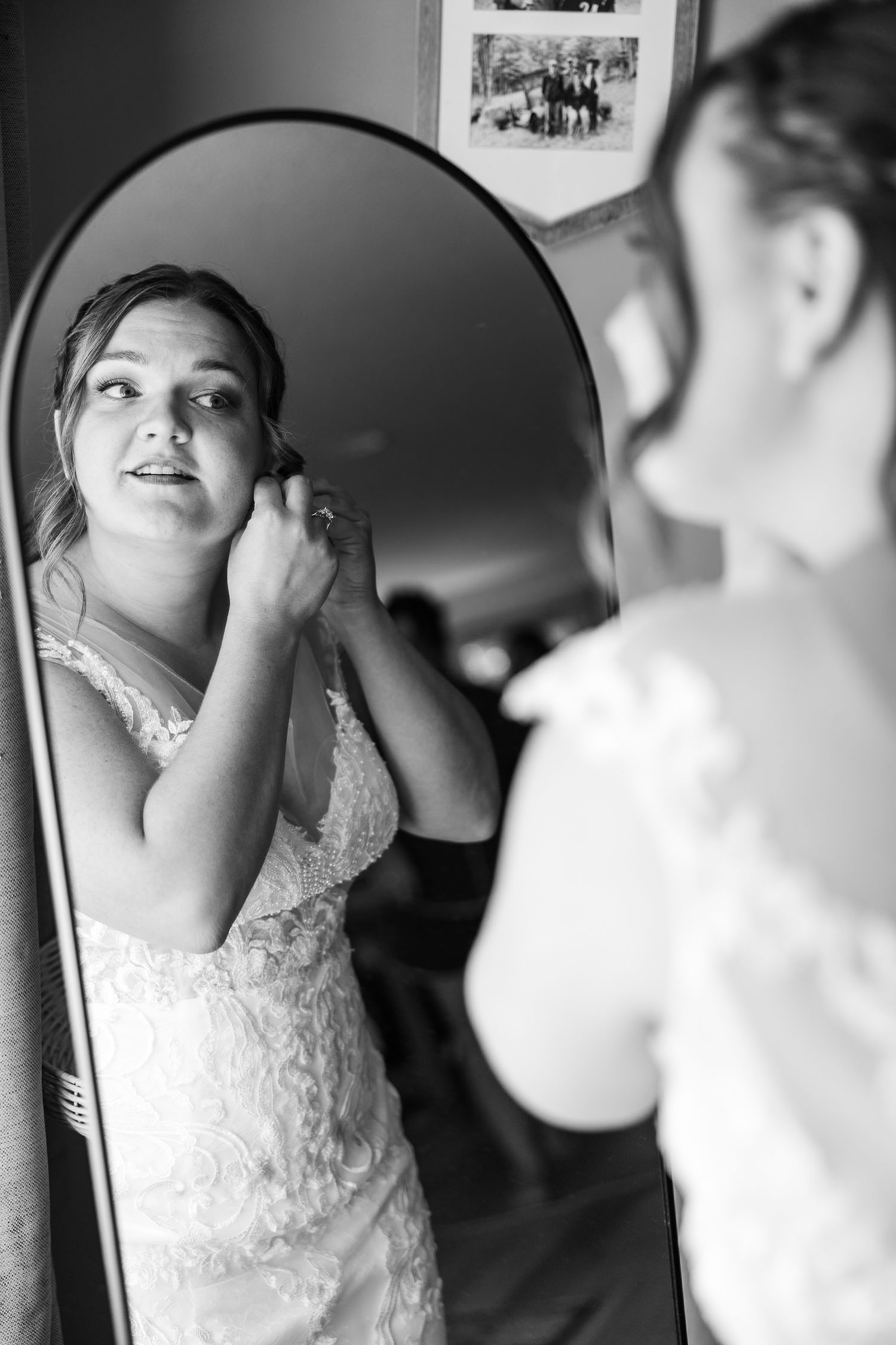 Bride in lace wedding gown adjusting earring while looking in arched mirror during wedding preparations.