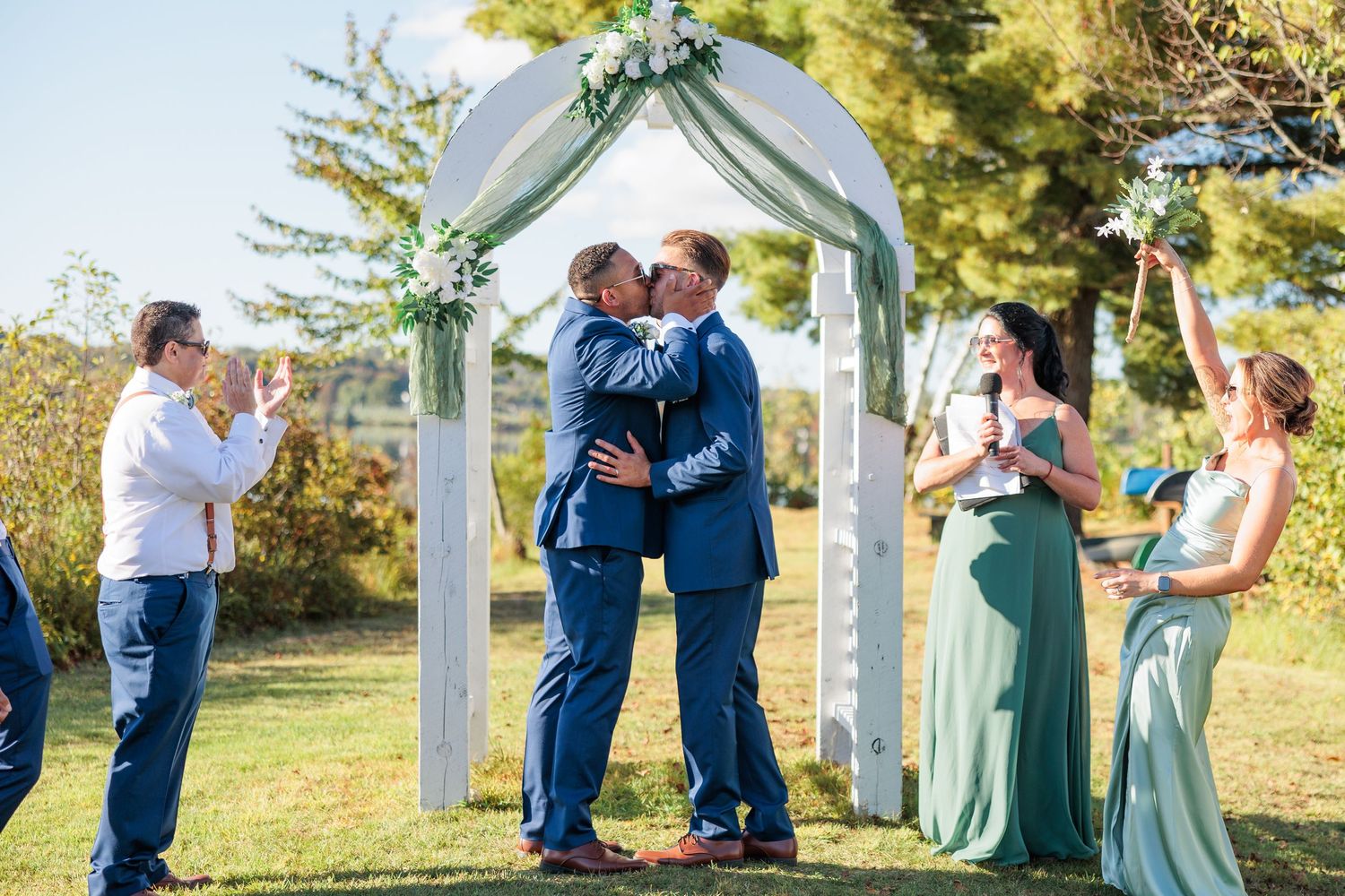 Wedding ceremony under white arch with sage green bridesmaid dresses and blue suits.