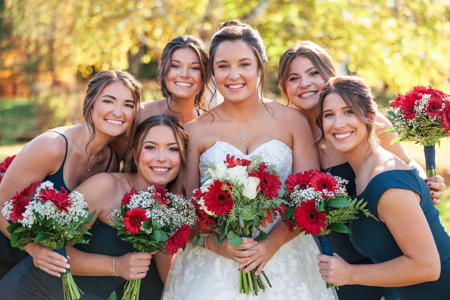 Wedding party members hold red and white bouquets while posing together on a sunny autumn day.
