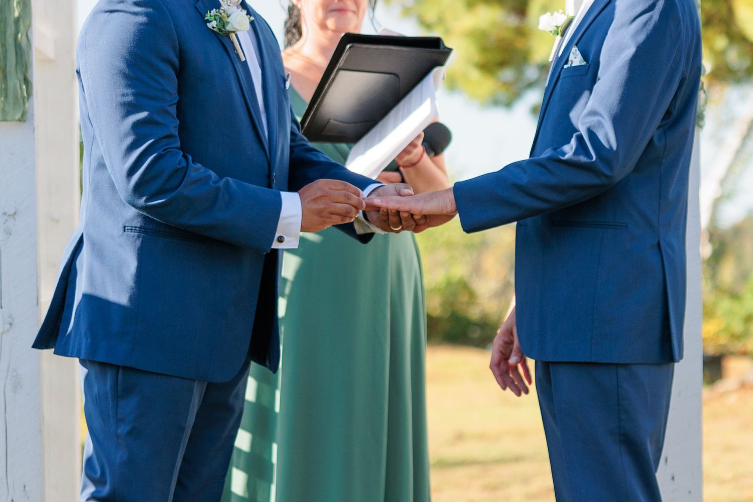 A couple in matching blue suits exchange vows under a white wedding arch decorated with greenery.