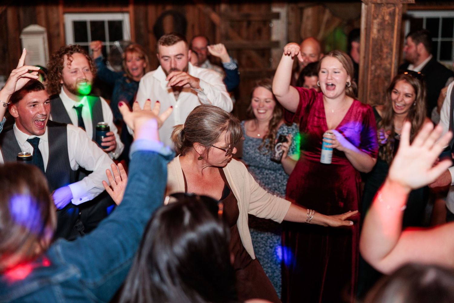 Wedding reception party in full swing with guests dancing under colorful lights in rustic barn venue.