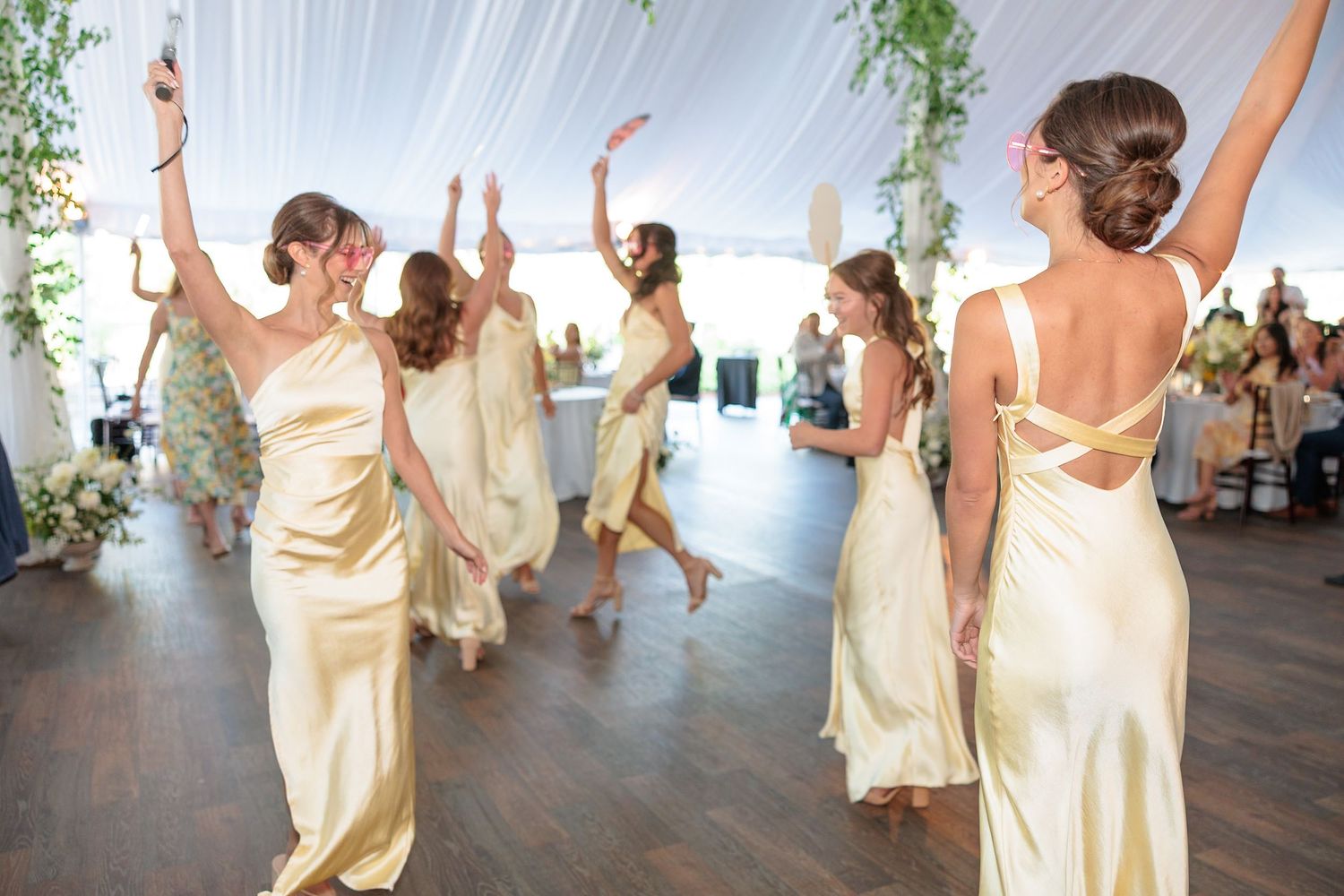 Bridesmaids in elegant gold satin dresses dancing joyfully with arms raised at outdoor tented wedding reception.