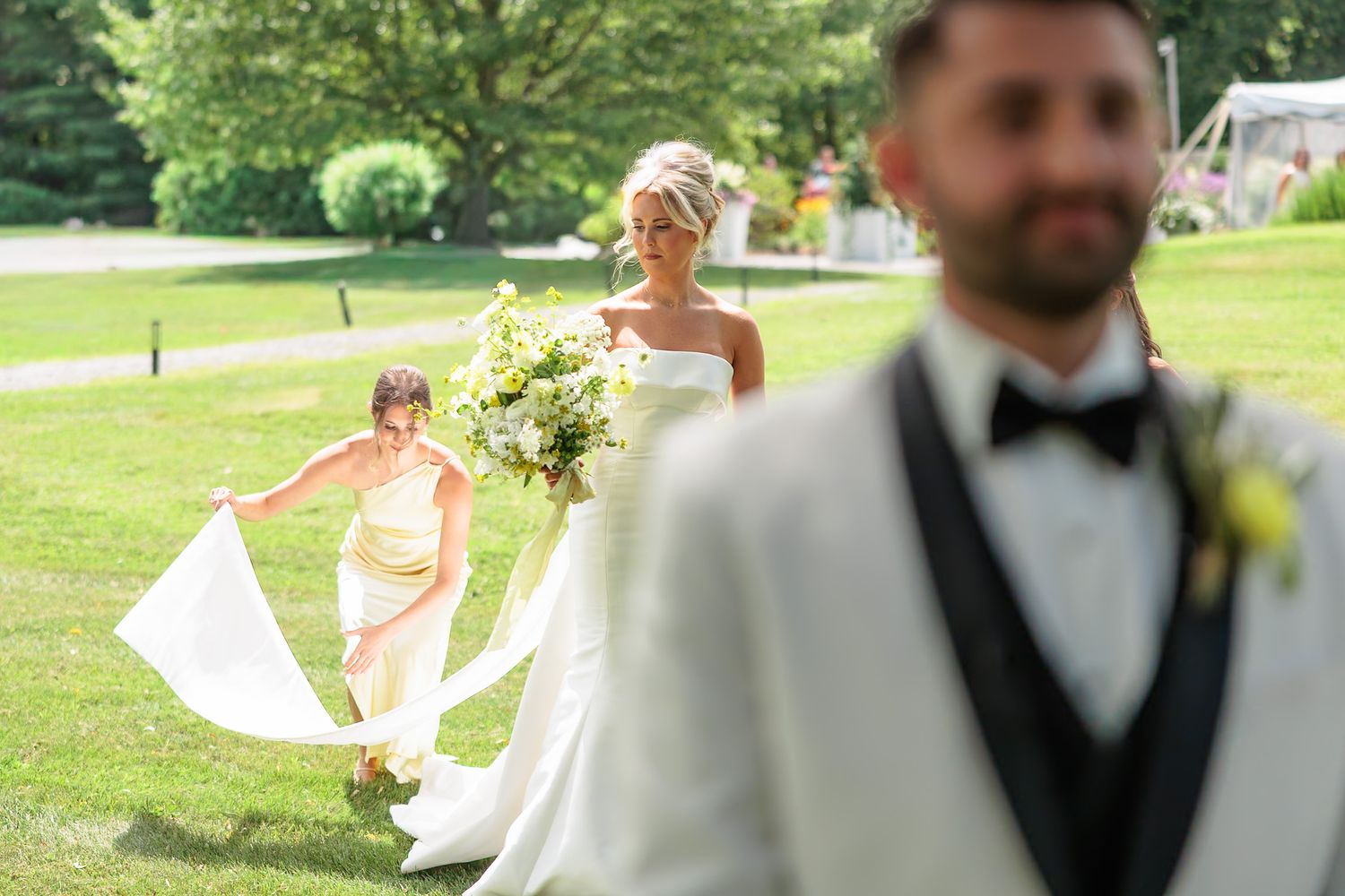 Bride in a white gown walking toward groom during first look moment assisted by bridesmaid in yellow dress.