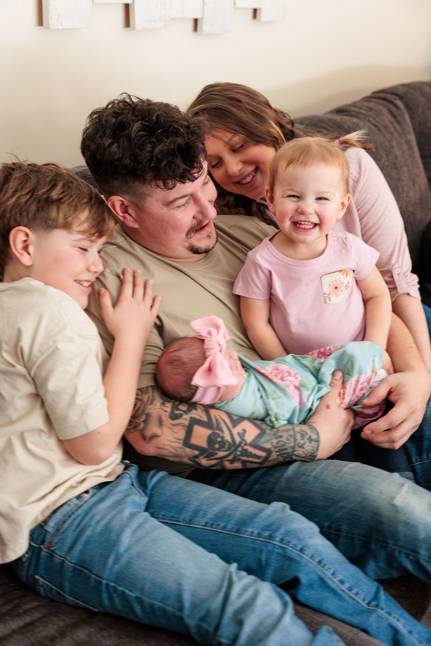 Smiling family with tattooed father holding newborn baby surrounded by toddler and young son on couch.