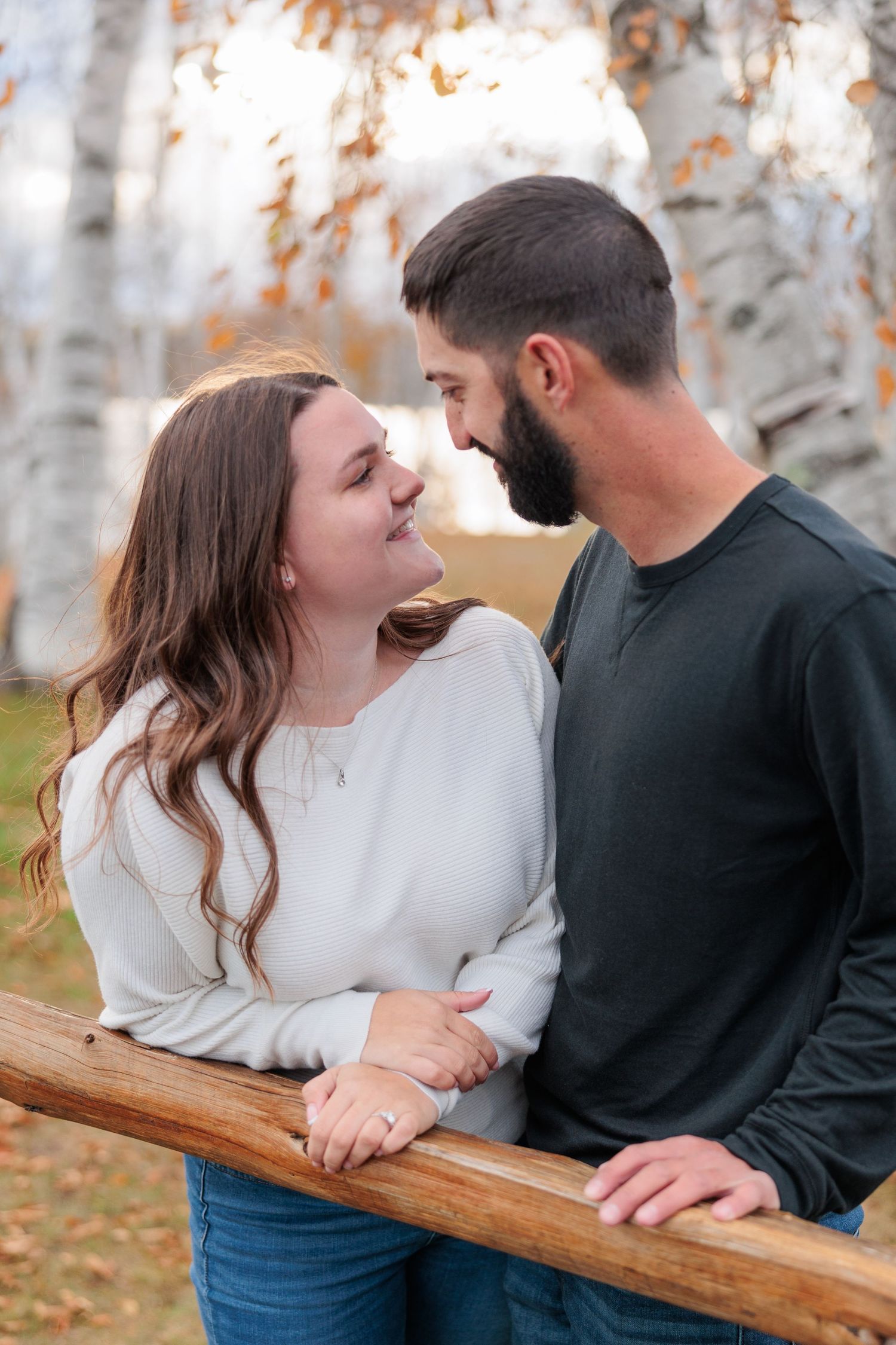 Romantic couple gazing into each others eyes near a wooden fence with birch trees in autumn background.