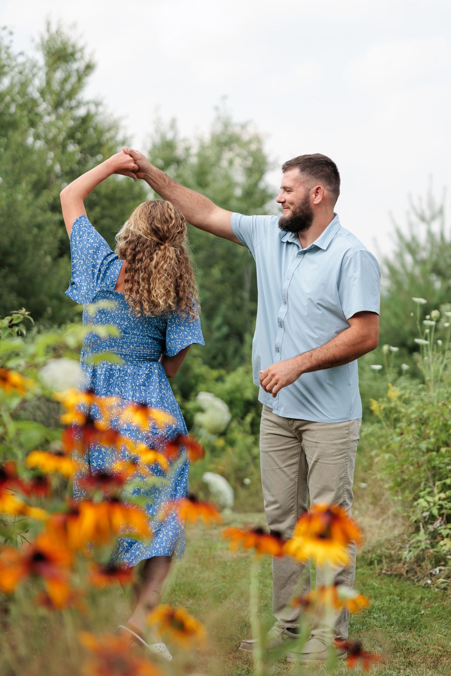 Bearded man spinning curly-haired woman in blue floral dress in lush summer garden with wildflowers.