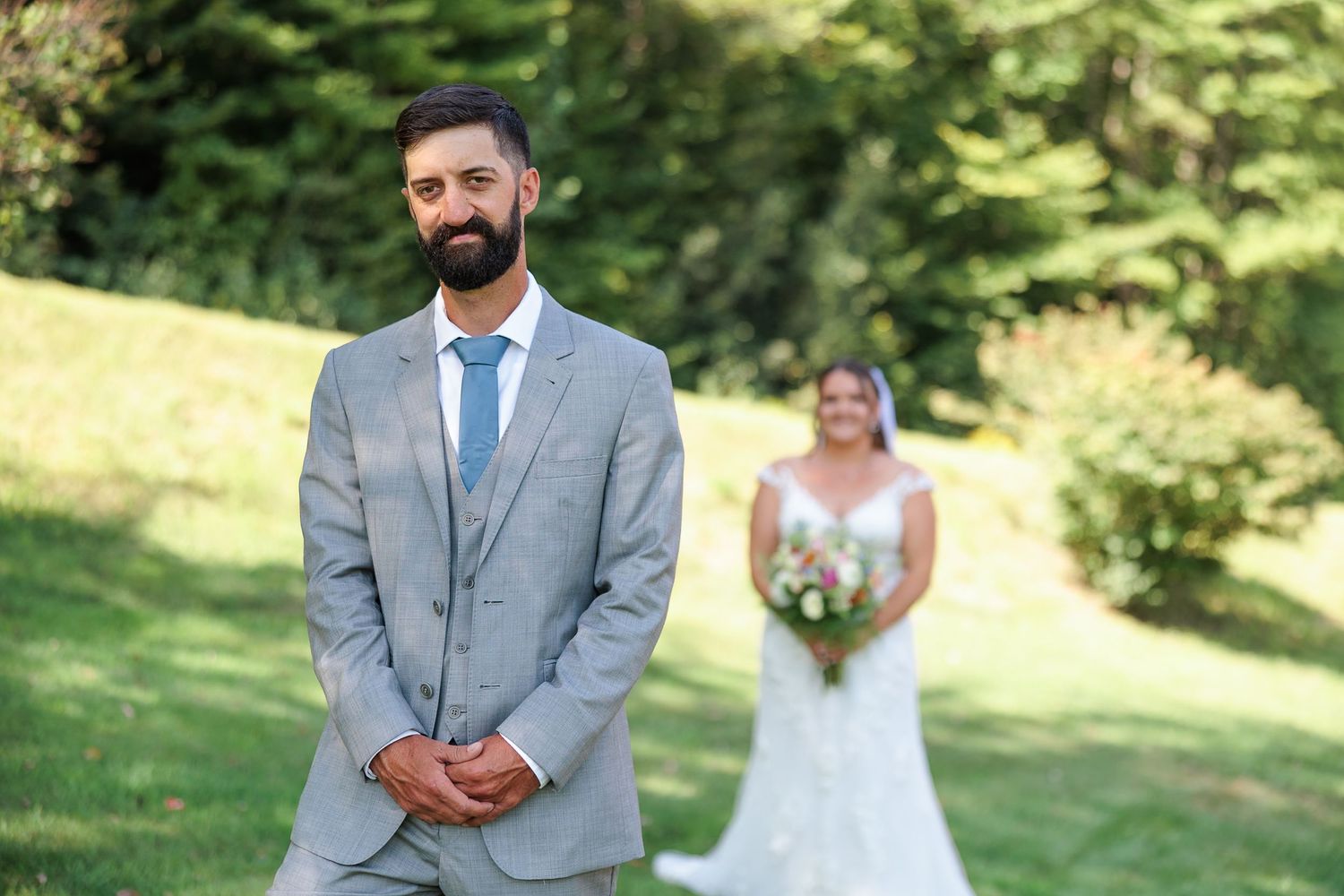 Groom stands in gray suit awaiting first look as bride approaches in background.