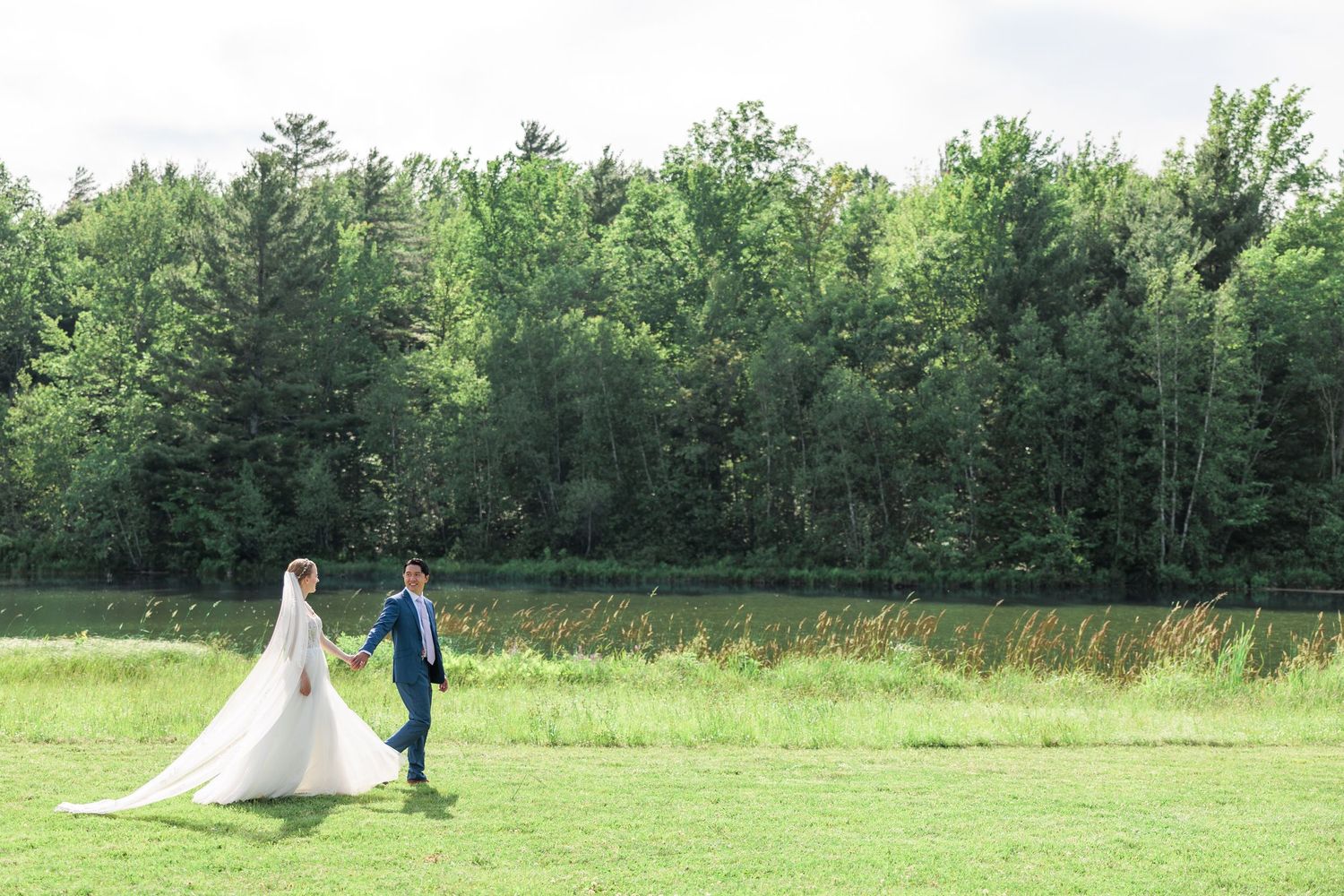 Bride in long white gown and groom in navy suit holding hands walking across a vast green field.
