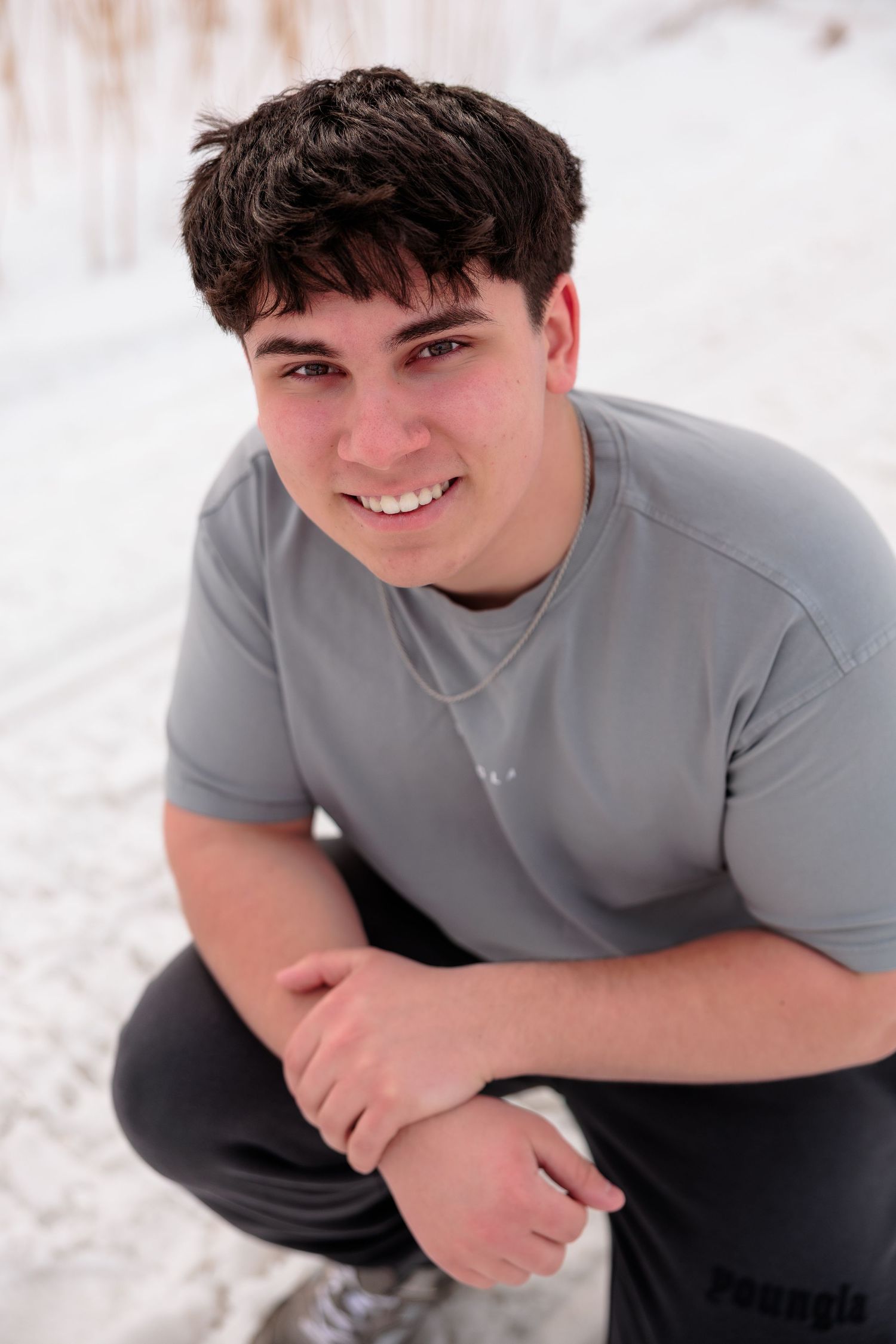 Smiling young man in gray t-shirt crouching in snow, wearing a necklace and dark pants outdoors.