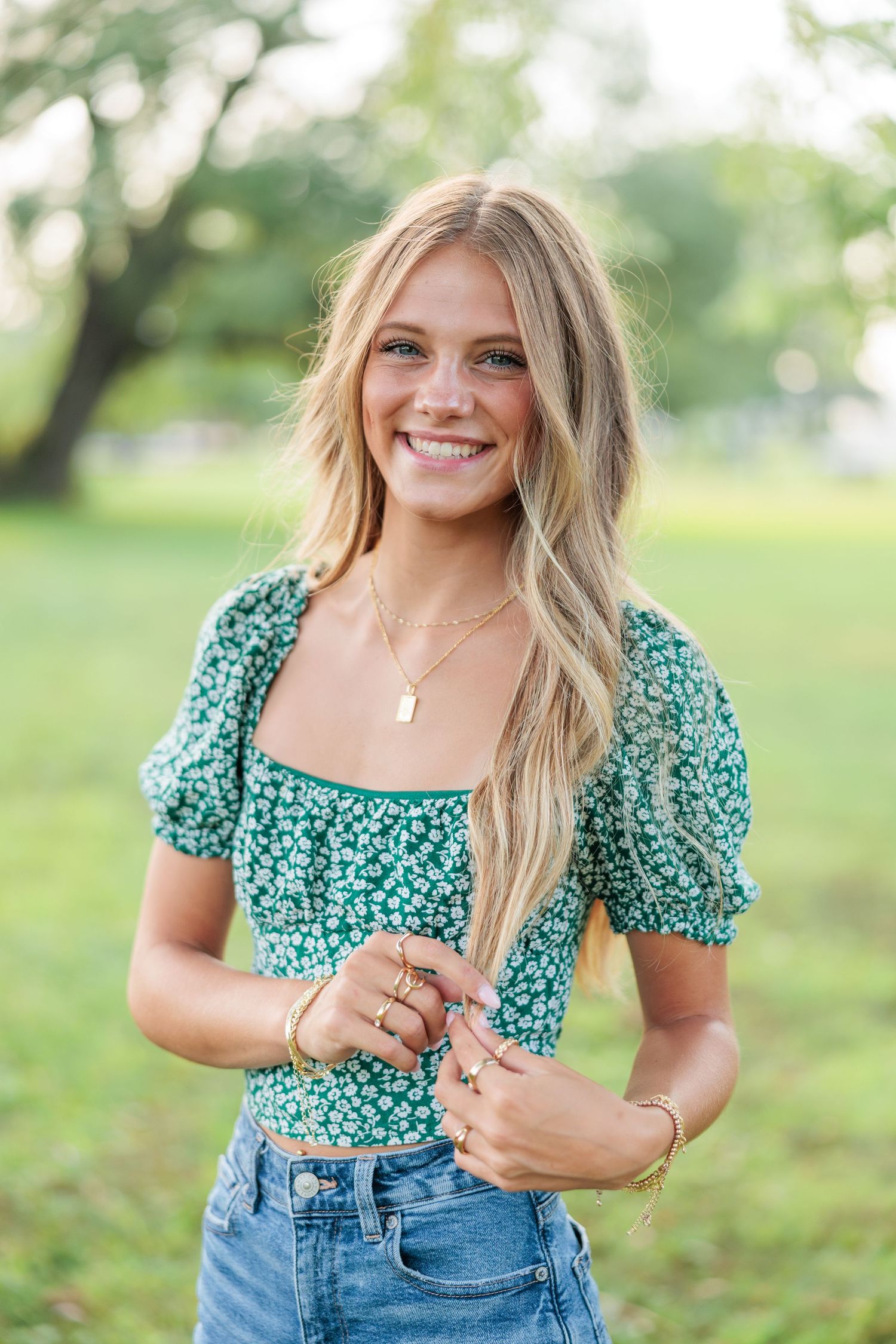 Young woman smiling outdoors in green floral crop top and jeans, with long blonde hair and gold jewelry.