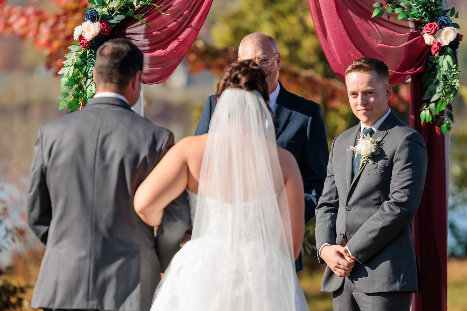 A couple exchanges vows during an outdoor wedding ceremony under a burgundy arch decorated with greenery.