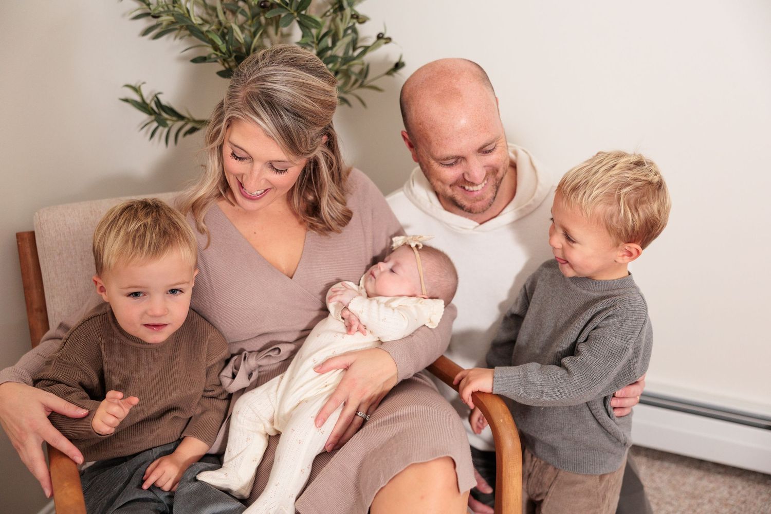 Family sits together on wooden chair sharing special moment with newborn baby in white outfit.