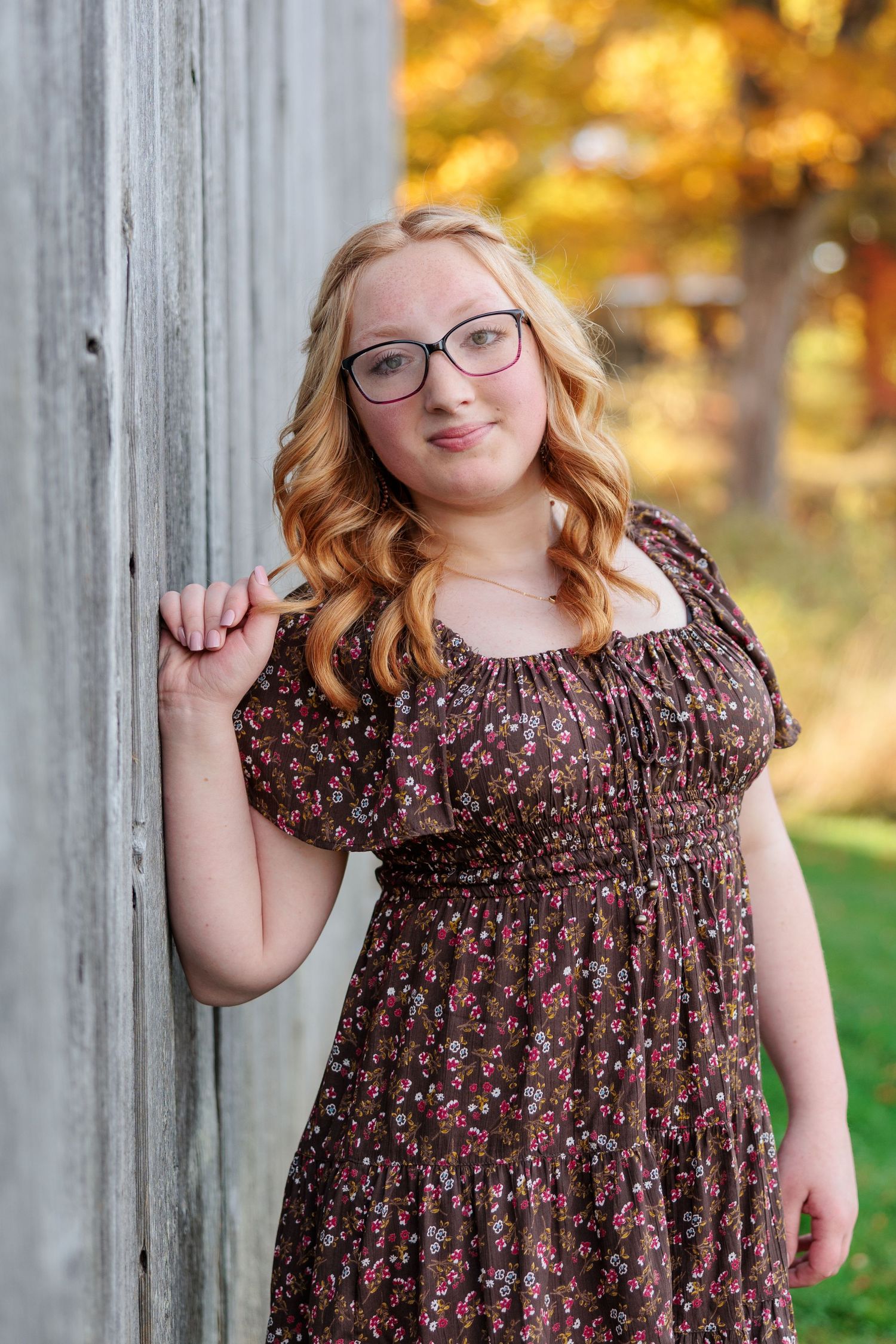 Young woman with blonde hair and glasses in floral dress leaning against rustic wood fence with autumn foliage background.