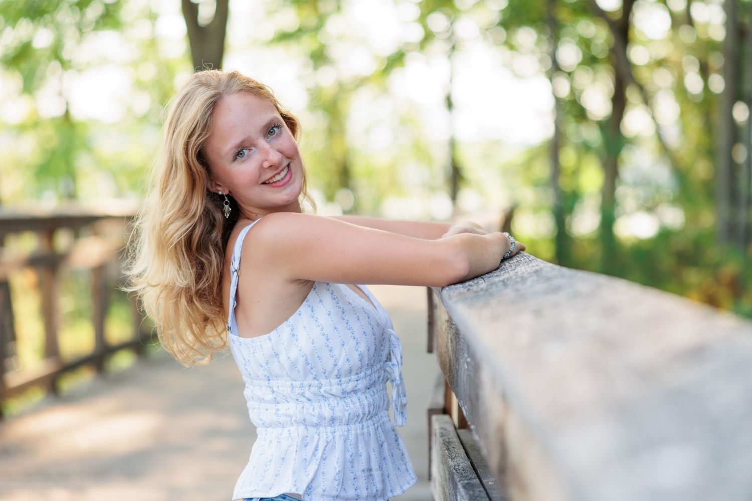 Young blonde woman in white top smiling outdoors, leaning on wooden railing surrounded by lush green trees.