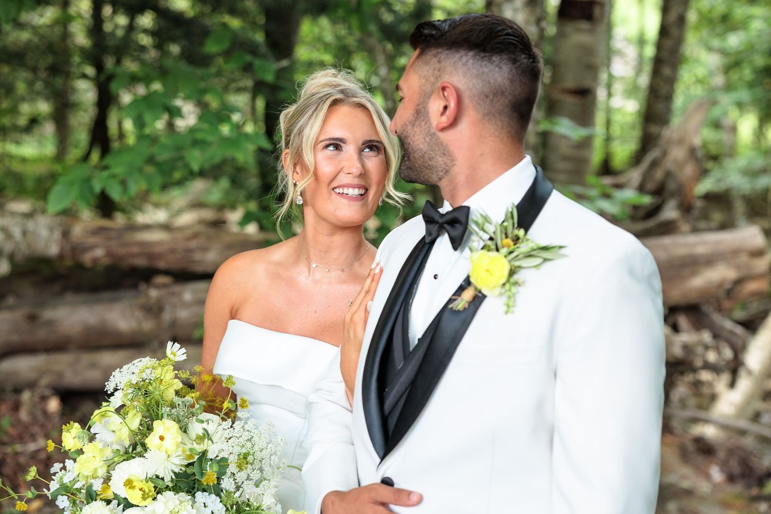 Bride and groom share a romantic moment in a lush forest, groom in white tuxedo with yellow boutonniere.