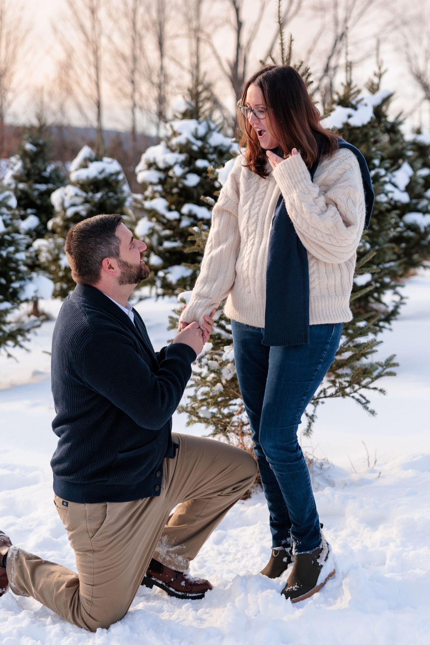 Man proposes on one knee to surprised woman in a snowy winter wonderland among snow-covered trees.