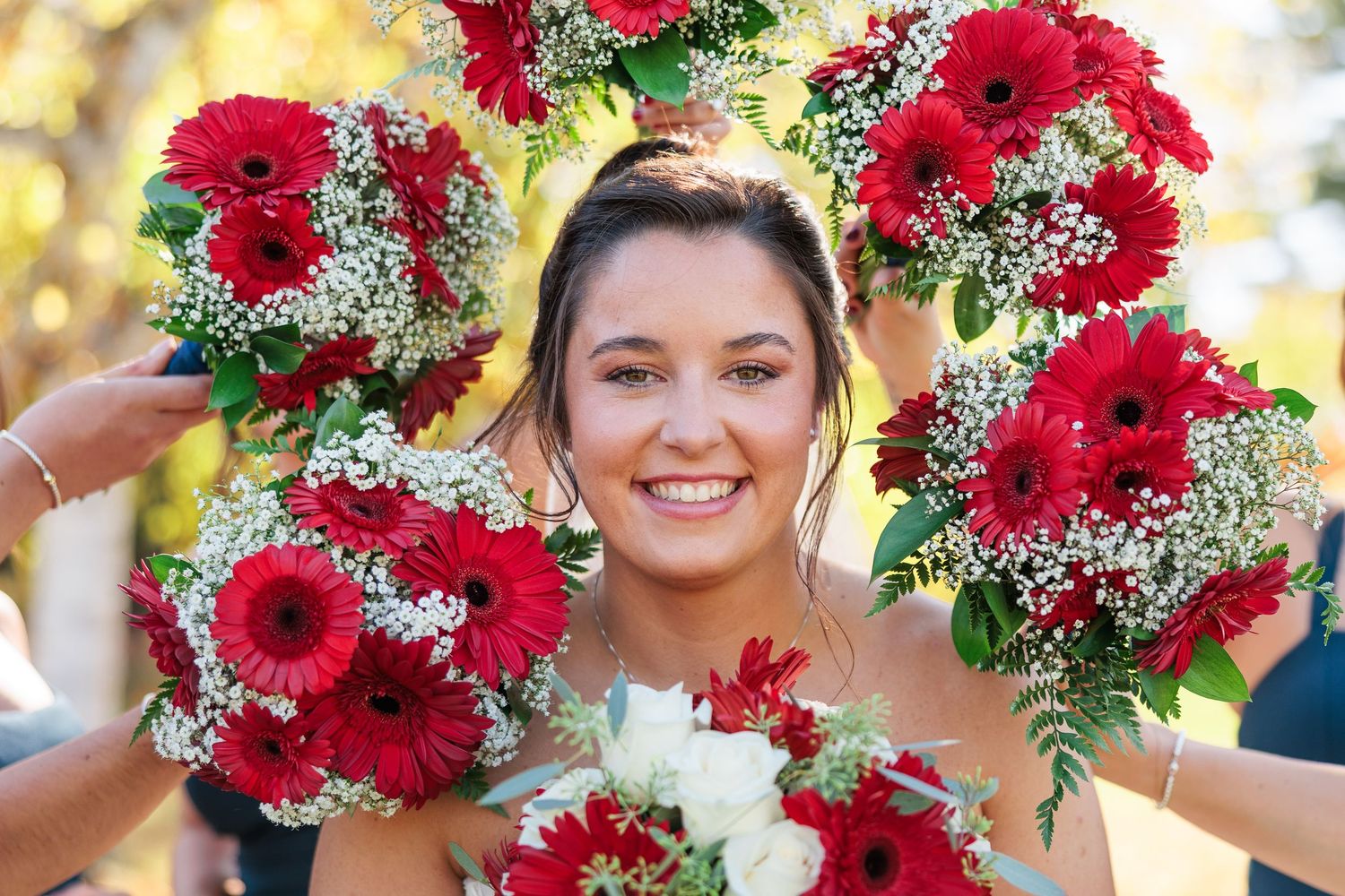 Smiling bride surrounded by red gerbera daisy and baby's breath bouquets held up by her bridesmaids outdoors.
