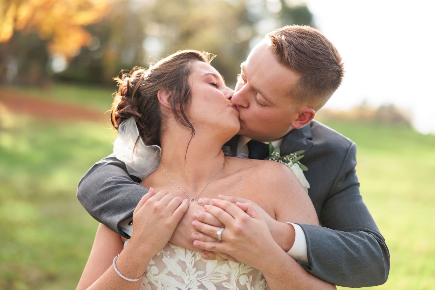 Groom embracing bride from behind as they share a romantic kiss outdoors at sunset.