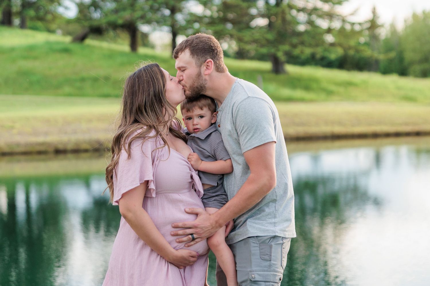 Pregnant woman in pink dress kissing partner while holding toddler son by a reflective pond outdoors.