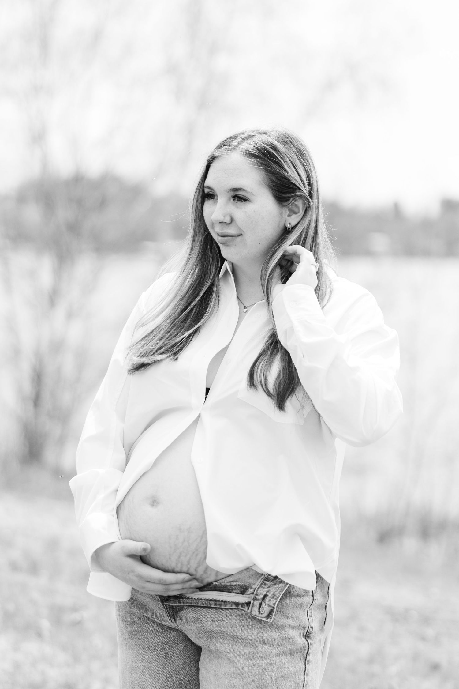 Black and white portrait of pregnant woman in open white shirt cradling baby bump outdoors near water.