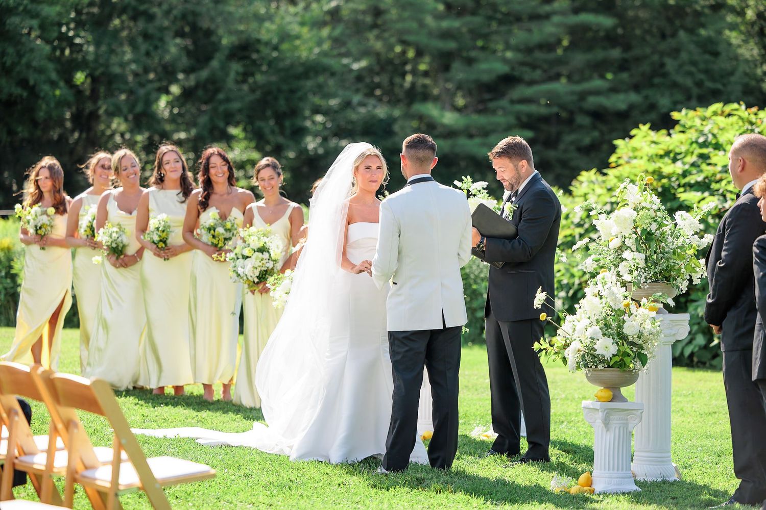 Bride and groom stand at outdoor altar with bridesmaids in pale yellow dresses during garden ceremony.