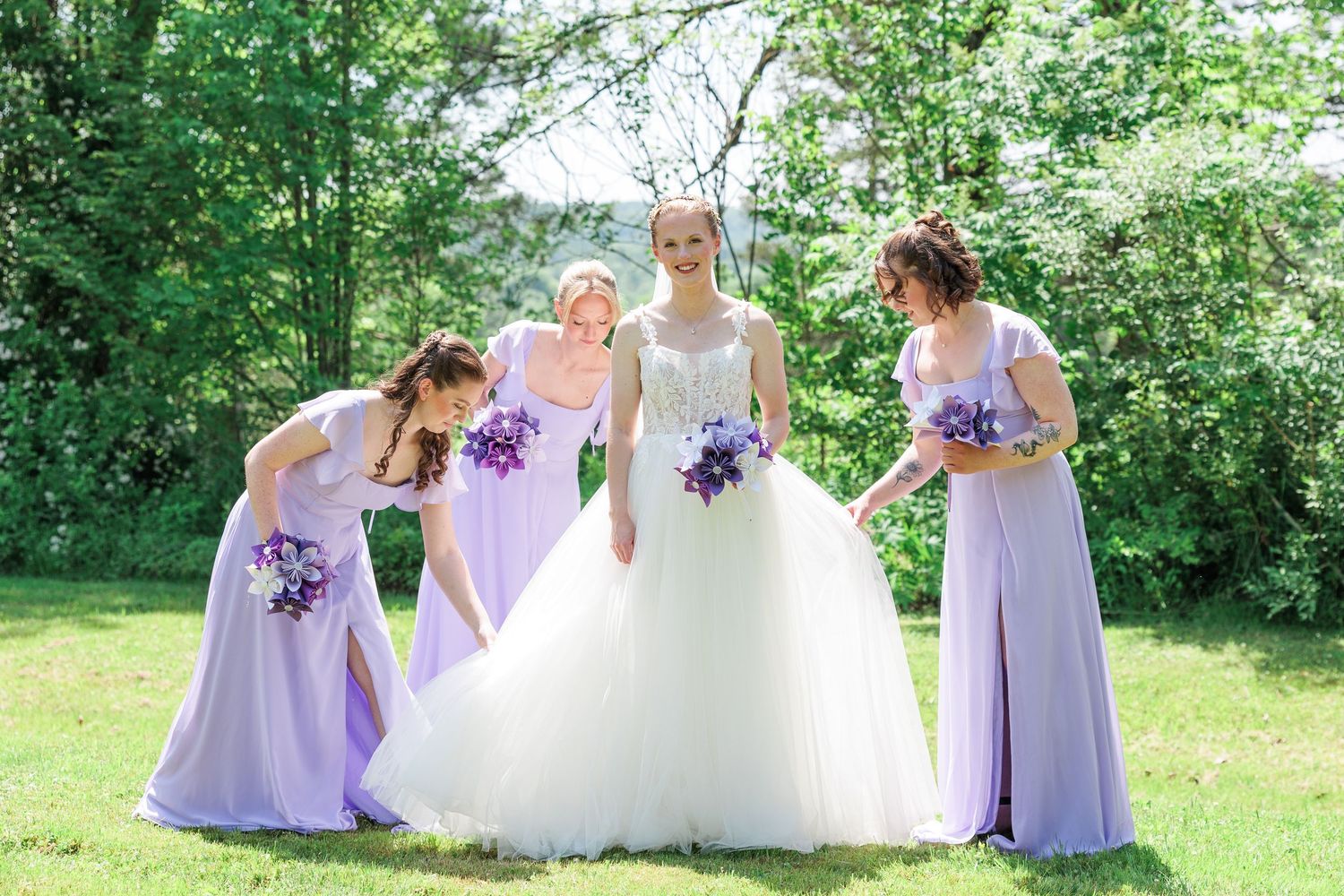 Bride in white ball gown with three bridesmaids in lavender dresses on green lawn.