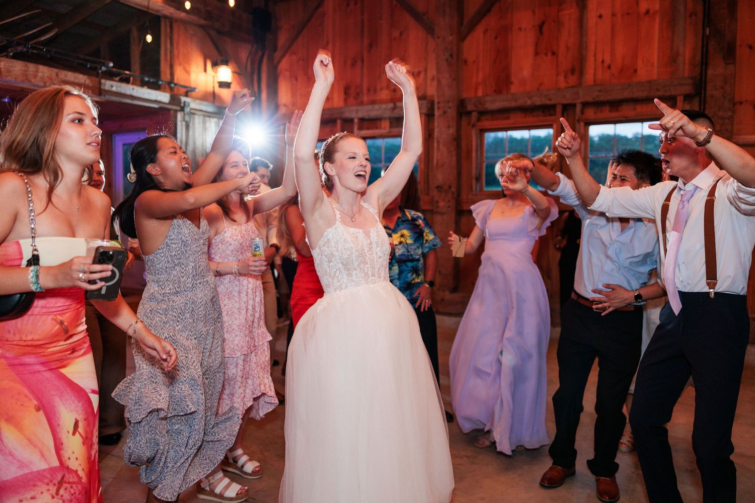 Bride in white gown celebrates with arms raised surrounded by cheering guests on the wedding dance floor.