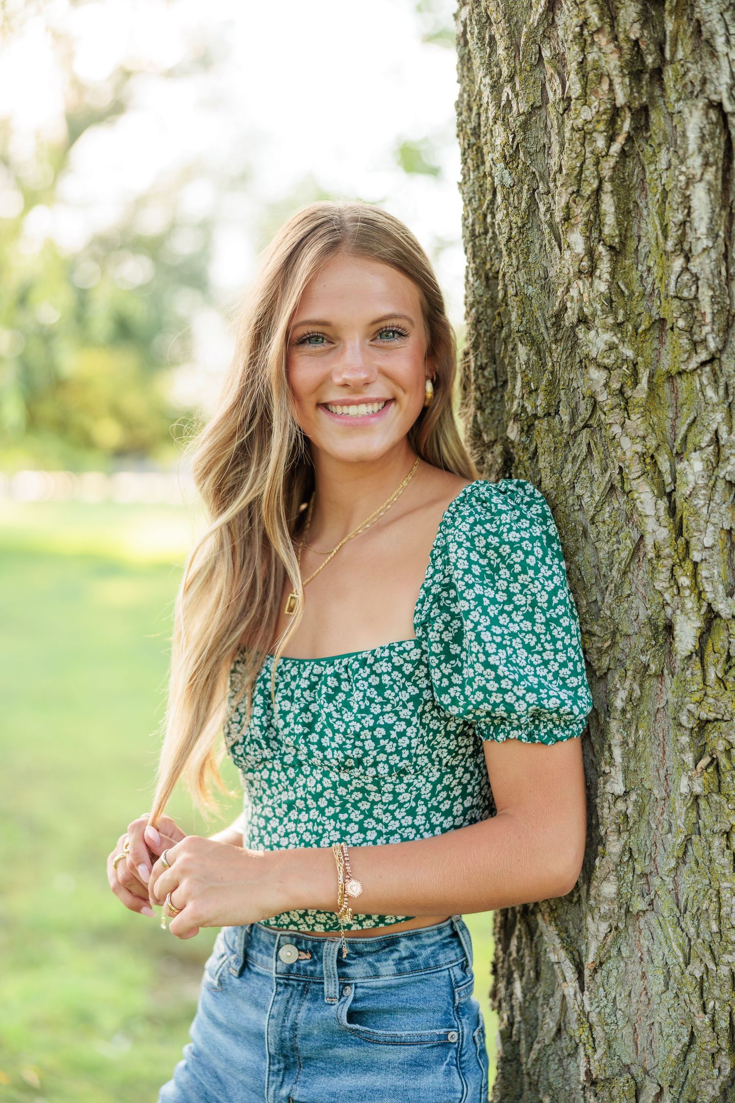 Smiling teenage girl in green floral crop top leaning against a tree in a sunny park.