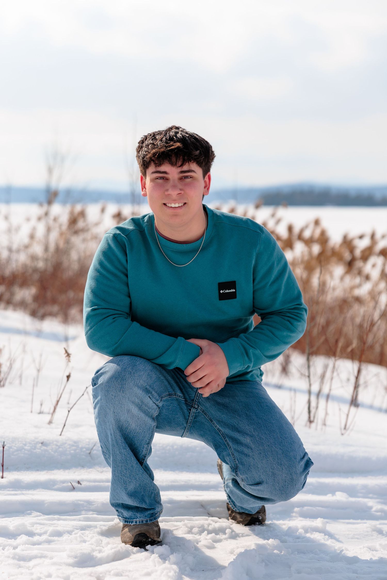 Young man in teal Columbia sweatshirt and jeans crouching in snow, smiling outdoors in winter landscape.