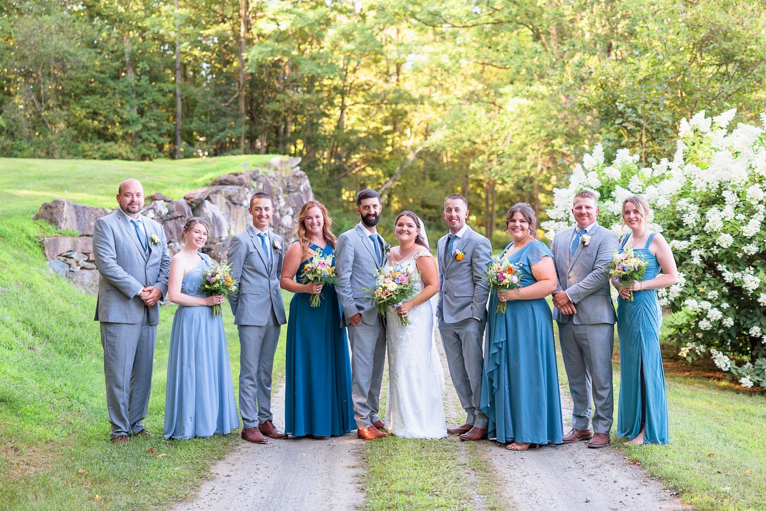 Wedding party poses on gravel path, bridesmaids in blue gowns and groomsmen in gray suits with lush greenery backdrop.