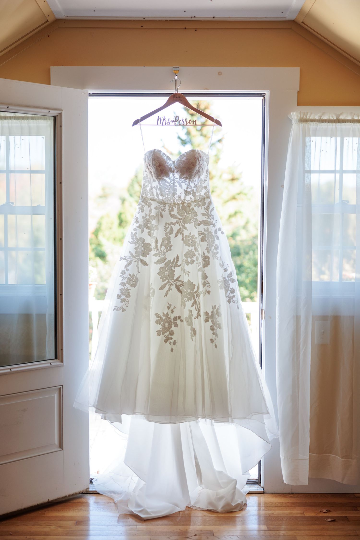 Elegant floral lace A-line wedding gown hanging in doorway on personalized Mrs. hanger with natural light.
