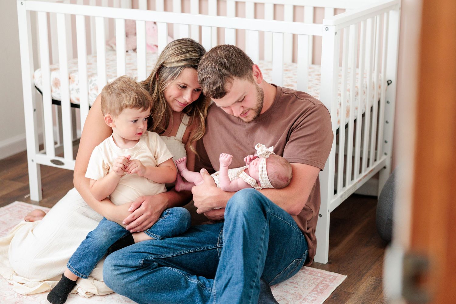 Family of four sitting together on nursery floor with toddler son and newborn baby girl.