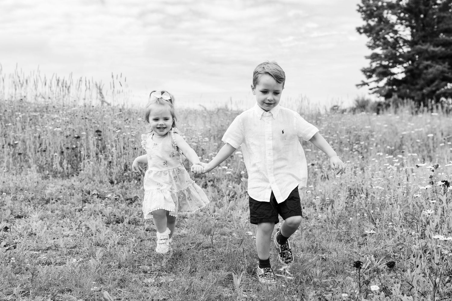 Black and white photo of two young children holding hands and running through a wildflower meadow.