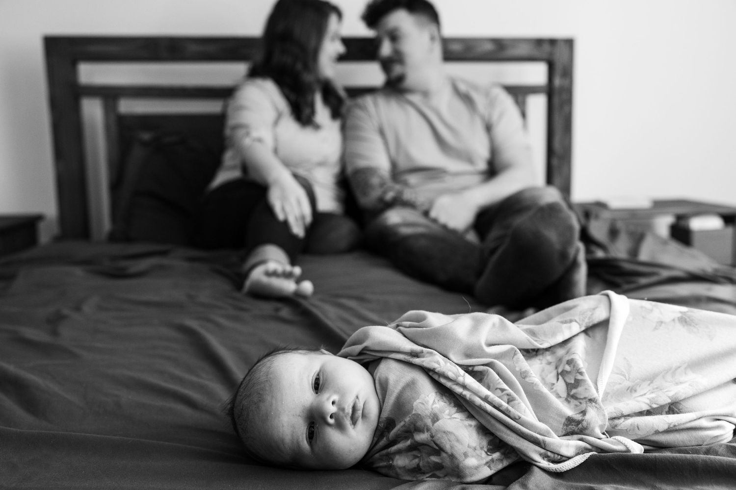 Black and white photo of parents relaxing on bed while newborn baby lies in foreground.