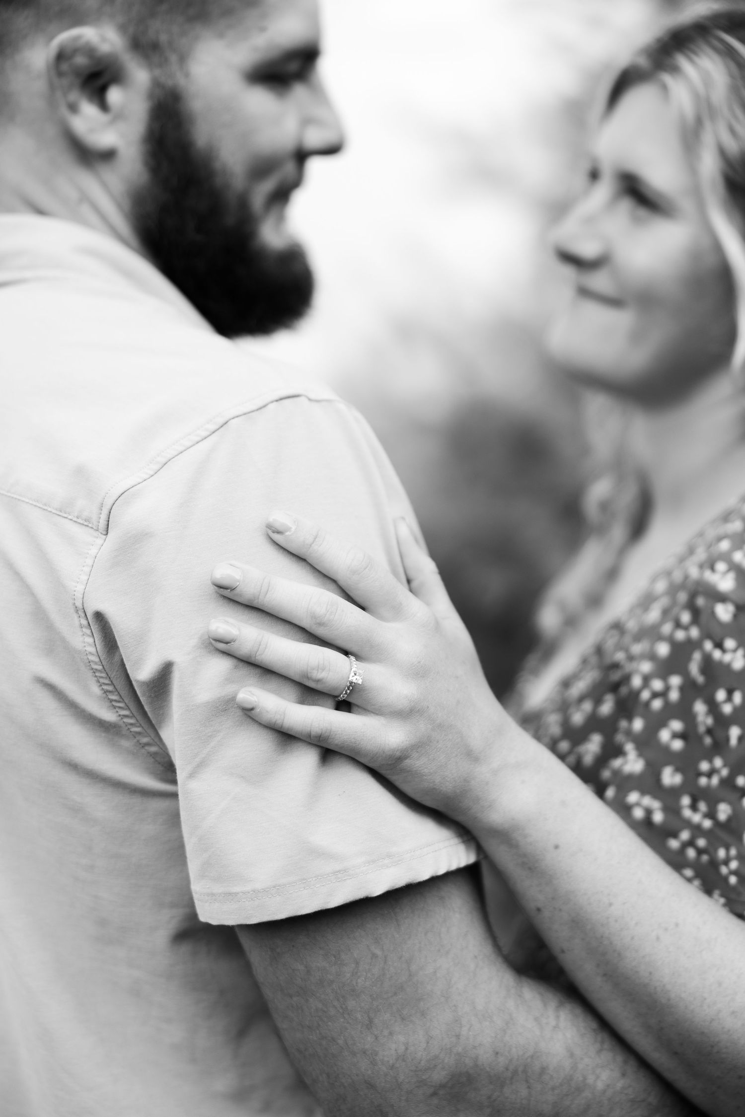 Black and white close-up of woman resting hand on mans shoulder showing engagement ring outdoors.