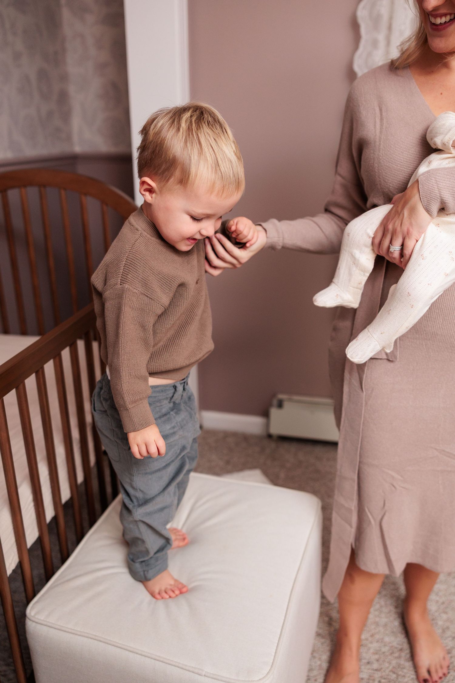 A toddler in a brown sweater stands on a cushion next to a wooden crib in a nursery.
