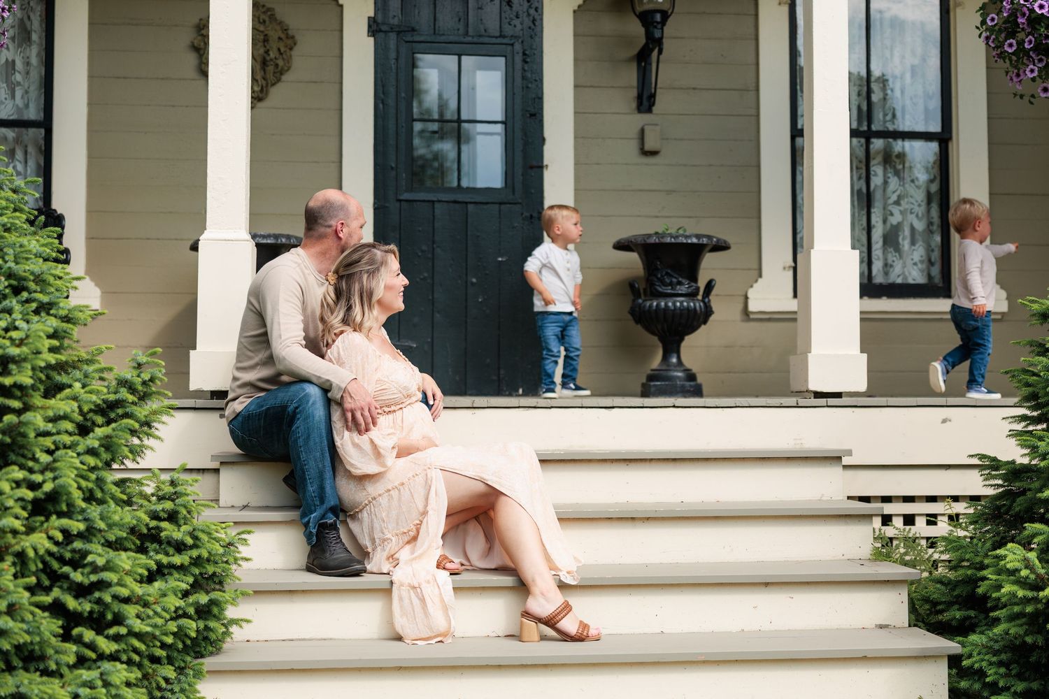 Couple sitting on porch steps while two children play behind them during maternity session.