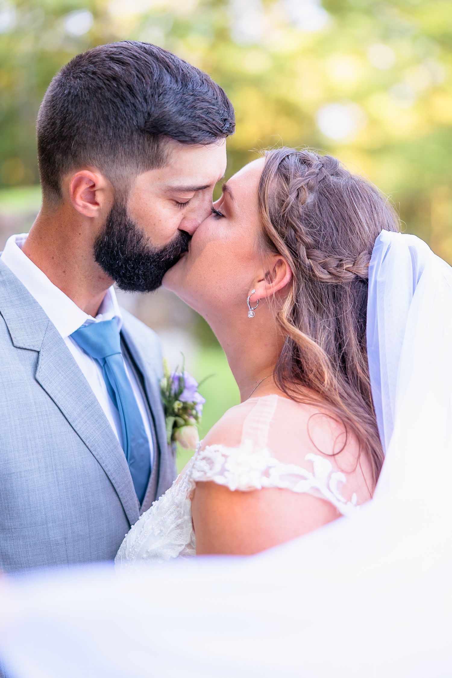 Bride and groom share a tender kiss outdoors, with the groom in a gray suit and blue tie at their wedding.