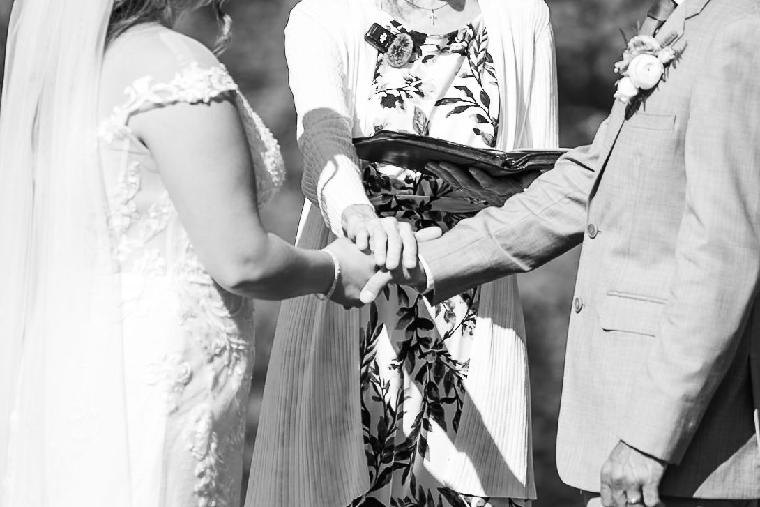 Officiant joins hands of bride and groom during intimate outdoor wedding ceremony in black and white photo.