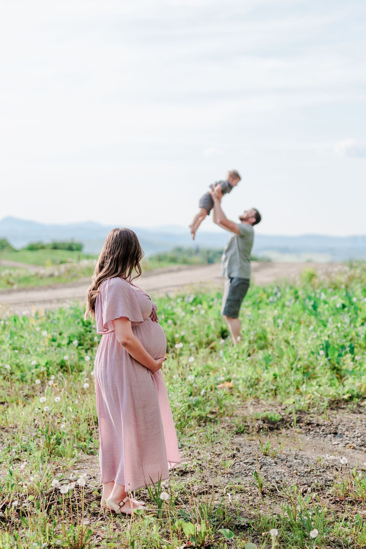 Pregnant woman watches husband toss toddler in the air in a green field.